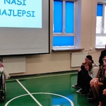 Uczniowie siedzący na ławeczkach słuchają koleżanki śpiewającej do mikrofonu.