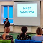 Pani pedagog mówi do mikrofonu. Wokół  siedzą uczniowie.