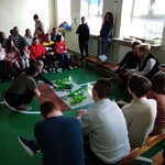 Warsztaty leśne - uczniowie siedzą na ławeczkach Po środku leży plansza z modelem lasu..