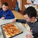 Dwóch uczniów siedzi przy stole. Jeden smaruje spód pizzy leżący na blaszce keczupem.