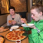 Nauczycielka i uczeń jedzą pizze. Przed nimi stoi duża pizza.