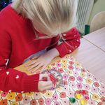 Uczennica siedzi przy stoliku i koloruje farbami bombkę.