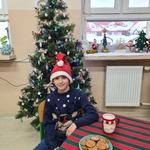 Uczeń w świątecznym sweterku i czapce mikołaja na głowie siedzi przy stole na którym stoi talerz z ciasteczkami i kubek z mlekiem.