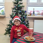 Uczeń w świątecznym sweterku i czapce mikołaja na głowie siedzi przy stole na którym stoi talerz z ciasteczkami i kubek z mlekiem.