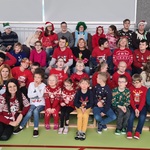 Na sali gimnastycznej siedzą na trybunach, w czterech rzędach, pani Dyrektor, nauczyciele oraz uczniowie. Wszyscy ubrani są w kolorowe, świąteczne sweterki. Część osób ma ma głowie czapki Mikołaja lub opaski świąteczne.