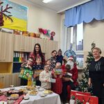 W rogu sali, z boku stołu przygotowanego do wigilii stoją uczniowie i uczennice klas pierwszych wraz z nauczycielkami i pomocami nauczyciela oraz Mikołajem. Wszyscy są odświętnie ubrani. Dzieci trzymają w rękach prezenty, które dostali od Mikołaja. Obok znajduje się pięknie ubrana choinka.