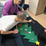 Uczniowie siedzą na podłodze i przyklejają świąteczne nalepki na zieloną papierową choinkę.