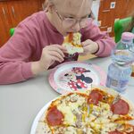 Uczennica siedzi przy stoliku i je kawałek pizzy. Przed nią stoi talerz z pizzą.