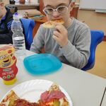 Uczeń siedzi przy stoliku i je kawałek pizzy. Przed nim stoi talerz z pizzą.