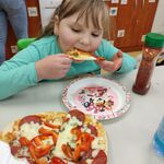 Uczennica siedzi przy stoliku i je kawałek pizzy. Przed nią stoi talerz z pizzą.