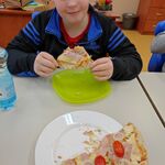 Uczeń siedzi przy stoliku i je kawałek pizzy. Przed nim stoi talerz z pizzą.