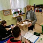 Pani Wicedyrektor trzyma w ręku pudełko z pączkami. Uczniowie siedzą przy stoliku,