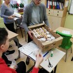 Pani Wicedyrektor trzyma pudełko z pączkami. Z tyłu stoi nauczycielka. Uczeń siedzi przy stoliku.