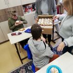 Pani Wicedyrektor trzyma w ręku pudełko z pączkami. Uczniowie siedzą przy stoliku,