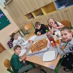 Uczniowie siedzą wokół stołu na którym znajduje się pudełko z dużą pizzą.