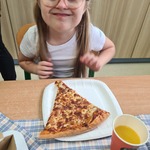 Uczennica siedzi przy stoliku. Przed nią na stole stoi talerz z kawałkiem pizzy i kubek z piciem.