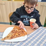 Uczeń siedzi przy stoliku i bierze kawałek pizzy z talerzyka.