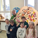 Przy ścianie, przed oknem klasowym stoi piątka uczniów z nauczycielką. Pani trzyma w rękach dużą pizzę wykonaną przez dzieci na szarym papierze i ozdobioną ,,dodatkami