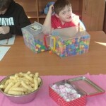 Uczeń siedzi przy stole. Przed nim stoją pudełka z klockami, miska z chrupkami i metalowe pudełko ze słodyczami.
