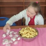Dziewczynka siedzi przy stole. Przed nią na stole stoi miska z chrupkami i leżą zapakowane w folie słodycze.