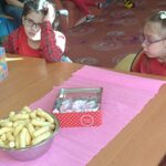 Uczennice siedzą przy stole. Przed nim na stole stoi pudełko ze słodyczami i miska z chrupkami.
