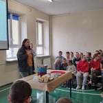 Uczniowie siedzący na ławkach słuchają opowieści pani prowadzącej warsztat.