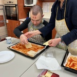 Uczeń z pomoca nauczyciela  kładzie plastry sera na spód pizzy posmarowany keczupem.