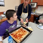 Uczeń kładzie plastry sera na spód pizzy posmarowany keczupem.