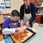 Uczeń kładzie plastry sera na spód pizzy posmarowany keczupem.