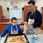 Uczeń z pomocą nauczyciela kładzie plastry sera na spód pizzy posmarowany keczupem.