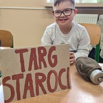 Uczeń siedzi przy stole na którym stoi tabliczka z napisem TARGI STAROCI I LEŻY stara żołnierska  menażka.