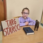 Uczennica siedzi przy stole na którym stoi tabliczka z napisem TARGI STAROCI i leży stara  książka.