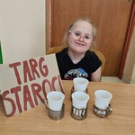 Uczennica siedzi przy stole na którym stoi tabliczka z napisem TARGI STAROCI  oraz kubeczki w starych uchwytach do szklanek.