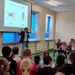 Uczniowie siedzą na ławeczkach i słuchają pana profesora omawiającego slajd wyświetlany na ekranie.