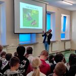Uczniowie siedzą na ławeczkach i słuchają pana profesora omawiającego slajd wyświetlany na ekranie.