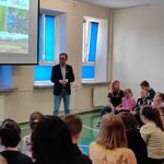 Uczniowie siedzą na ławeczkach i słuchają pani dyrektor mówiącej do mikrofonu.