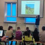 Uczniowie siedzą na ławeczkach i słuchają pana profesora omawiającego slajd wyświetlany na ekranie.
