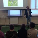 Uczniowie siedzą na ławeczkach i słuchają pana profesora omawiającego slajd wyświetlany na ekranie.
