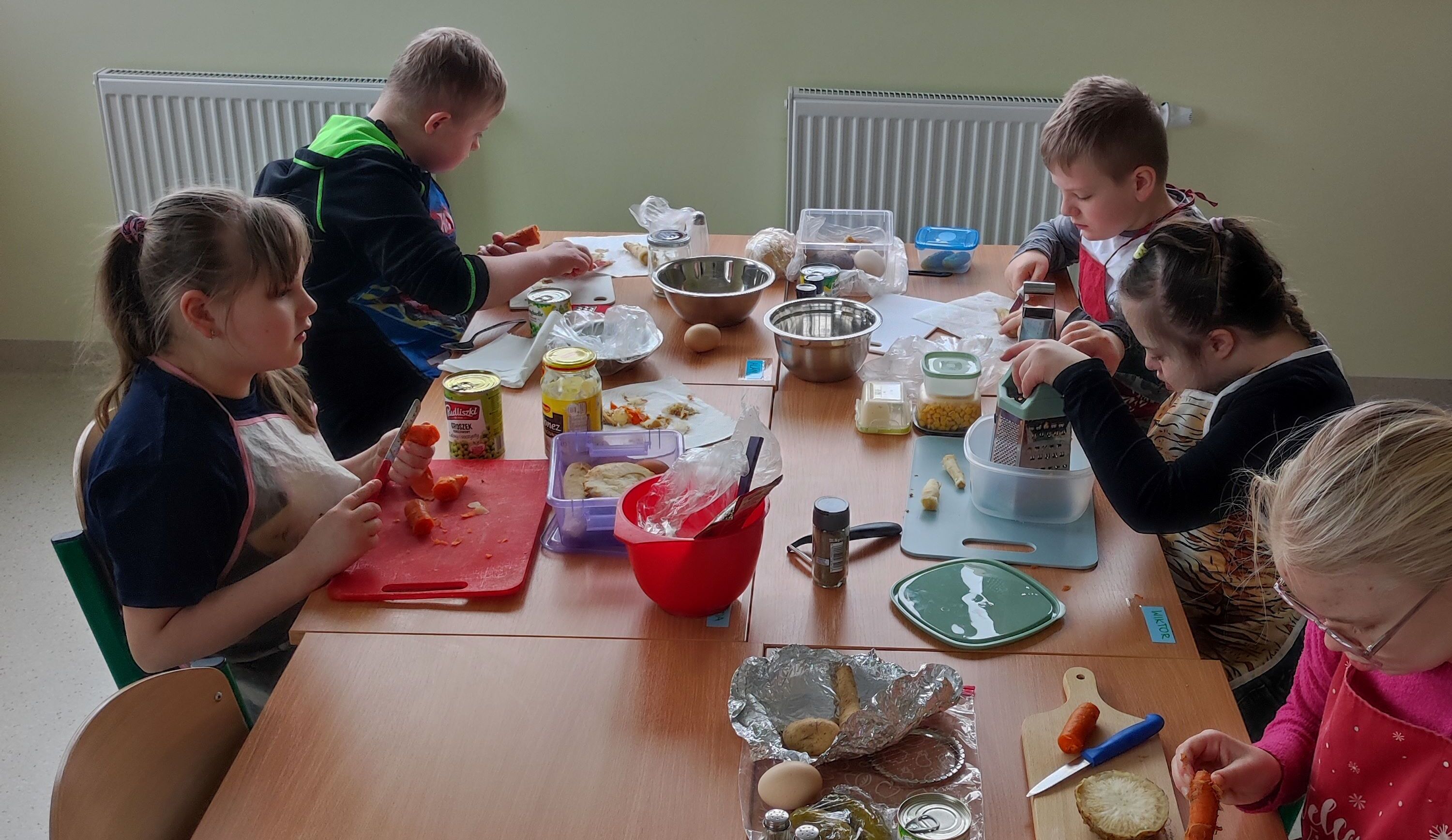 Uczniowie siedzą wokół stołu. przed nimi na stole leżą produkty spożywcze do przygotowania sałatki.