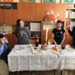 Uczennice stoją wokół stołu i śpiewają sto lat jubilatce.