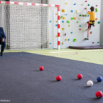Pan prezydent Białegostoku Tadeusz Truskolaski rzuca kulą do gry w bocce. W tle nauczyciel i uczeń w trakcie zajęć na ściance wspinaczkowej.