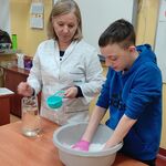 Nauczycielka z miarką w ręku przygląda się uczniowi mieszającemu mąkę z wodą w misce.