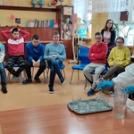 Uczniowei siedzą na krzesłach i przyglądają się nauczycielce wsypującej barwnik do szklanki z wodą.