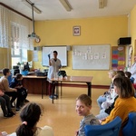 Uczniowie siedzą na krzesłach i obserwują nauczycielkę w białym fartuchu wykonującą eksperyment.