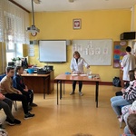 Uczniowie siedzą na krzesłach i obserwują nauczycielkę w białym fartuchu wykonującą eksperyment.