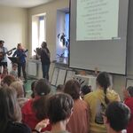 Uczniowie siedzą na ławkach i śpiewają piosenkę z paniami grającymi na gitarach.