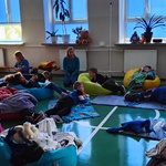 Uczniowie leżą na podłodze, pufach i karimatach przykryci kocami. Na ławce siedzą nauczycielki.