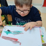 Uczeń siedzi przy stole i maluje pędzlem na dużej białej kartce. Przed nim stoi pudełko z farbami i pojemnik z wodą.