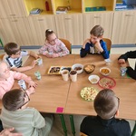 Uczniowie siedzą wokół stołu na którym stoją pojemniki z różnymi płatkami.