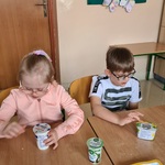 Uczennica  i uczeń siedzą przy stole. Przed nimi stoją pojemniki z jogurtem i serkiem homogenizowanym.